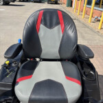 Front-facing view of a black and gray leather mobility scooter seat with red accents and joystick control on the left, in a parking lot.