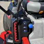 Close-up of a ride-on machine's cockpit: gray seats with red stitching, black control panel, joystick, yellow emergency stop button, and red gear selectors on the left.
