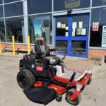 Red and black Toro riding lawn mower parked outside a glass-front building with blue doors and yellow bollows in front.