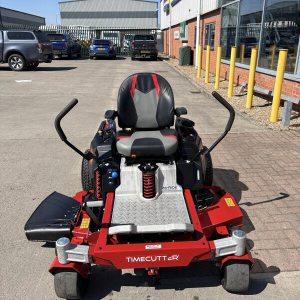 Red TimeCutter riding lawn mower in the foreground outside a commercial building with parked cars in the background.