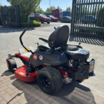 Red riding lawn mower in foreground with a black seat, parked on a paved area near a metal fence; a shelved parking lot with cars and a tree in the background.