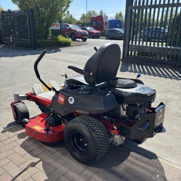 Red riding lawn mower in foreground with a black seat, parked on a paved area near a metal fence; a shelved parking lot with cars and a tree in the background.