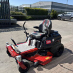 Red and black Toro ride-on lawn mower in a parking lot with industrial warehouses and a silver pickup in the background on a sunny day, blue sky above.
