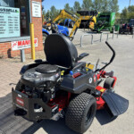 Riding lawn mower in the foreground outside an equipment shop, with tractors and machinery lined up in the lot behind.