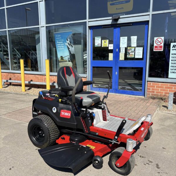 Red and black Toro riding lawn mower parked outside a glass-front building with blue doors and yellow bollows in front.