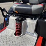 Close-up of a mobility device suspension area: red coil spring under a gray-black seat with red stitching, diamond-plate foot platform, and MYRIDE Suspension System badge visible on the side.