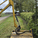Yellow excavator with bucket attached, on a grassy field beside a hedge and distant farm buildings under a blue sky.