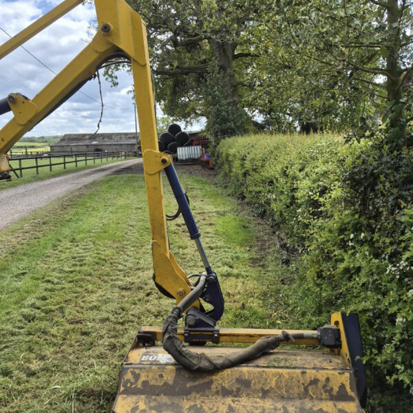 Yellow excavator with bucket attached, on a grassy field beside a hedge and distant farm buildings under a blue sky.