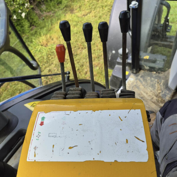 Close-up of multiple levers in a heavy machinery cab, with one orange lever among black ones, yellow control panel in foreground and greenery outside.
