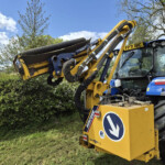Yellow construction excavator with a hydraulic lifting arm holding a large log against a blue sky, with surrounding trees and bushes nearby