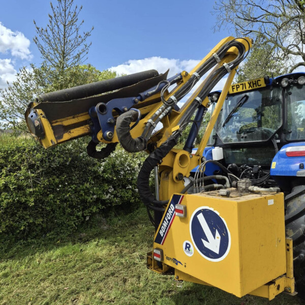 Yellow construction excavator with a hydraulic lifting arm holding a large log against a blue sky, with surrounding trees and bushes nearby