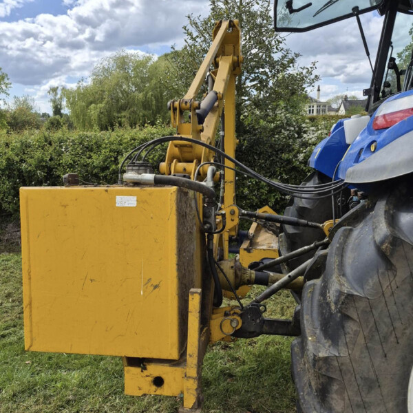 Yellow construction machine with hydraulic arm attached to a blue vehicle in a garden setting, with hedges and a cloudy sky behind it.