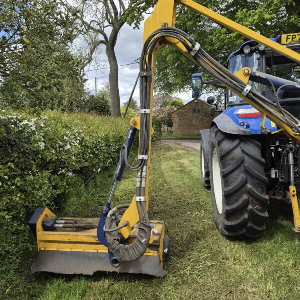 Blue tractor with a yellow hydraulic arm and hoses, operating a ground attachment in a grassy residential yard beside a hedge and trees.