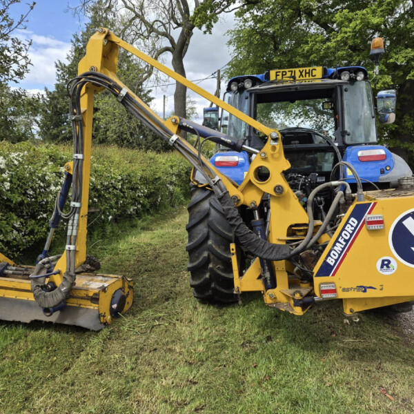 Yellow construction vehicle with a hydraulic arm parked on a grassy verge beside a hedge; blue cab and warning sign visible, likely at a roadside worksite.