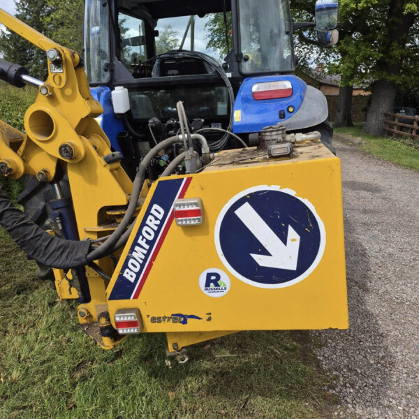 Back of a yellow construction vehicle with a blue cab, leaning road-sign in front on a gravel path outside grass and trees.