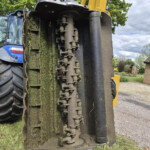 Close-up of a muddy excavator track and hydraulic components, with a yellow boom visible on a grassy site.