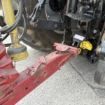 Close-up of a red agricultural implement attached to a tractor hitch, showing hydraulic hoses, metal linkages, and a worn, dirty frame.