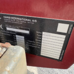 Close-up of a black identification plate on a red surface, reading HARDI INTERNATIONAL A/S and address information.