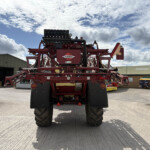 Rear view of a red HARDI agricultural sprayer with multiple nozzles on top, large tires, parked in an industrial yard under a cloudy sky.