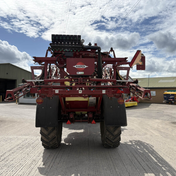 Rear view of a red HARDI agricultural sprayer with multiple nozzles on top, large tires, parked in an industrial yard under a cloudy sky.