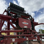 Red agricultural sprayer frame with black hoses, green valve blocks, and a white gauge against a blue sky; brand name HARDI visible on the side.