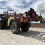 Red agricultural sprayer equipment mounted on a tractor, on display outdoors at a dealership.