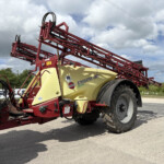 Red agricultural sprayer with a yellow tank and extended red booms, parked by a field under a cloudy sky.