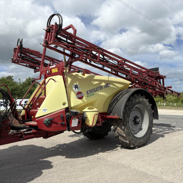 Red agricultural sprayer with a yellow tank and extended red booms, parked by a field under a cloudy sky.
