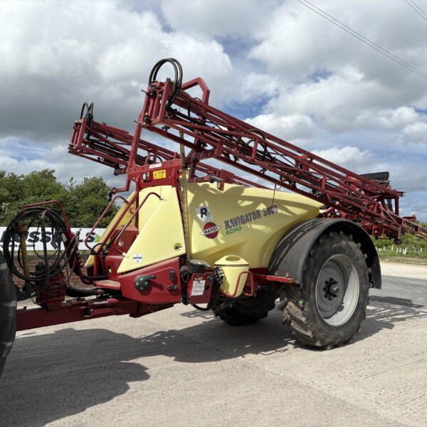 Red farm sprayer with a yellow tank and large tires parked on a paved area beneath cloudy skies, hoses and booms visible.