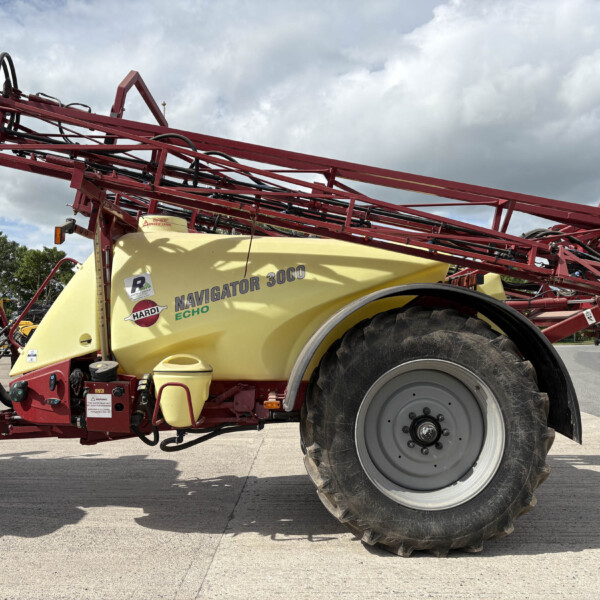 Red agricultural sprayer frame with a yellow Navigator 3000 tank, hoses and controls, parked on pavement under a cloudy sky.