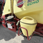 Close-up of a yellow agricultural sprayer tank with red frame, hoses, and warning labels visible on the equipment.