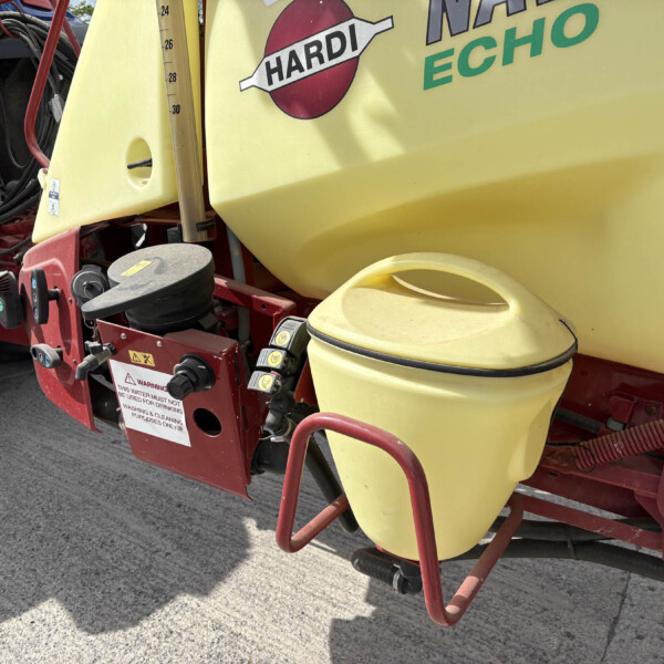 Close-up of a yellow agricultural sprayer tank with red frame, hoses, and warning labels visible on the equipment.