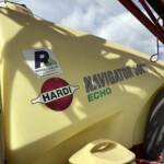 Close-up of a yellow agricultural sprayer with a vertical gauge on the left, branding stickers (HARDI, Russell Group Agriculture) and red frame parts above against a blue sky.