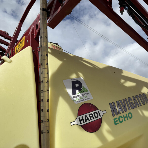 Close-up of yellow agricultural equipment with a red frame, a vertical gauge, and warning stickers including 'HARDI' and 'NAVIGATOR 3000' text visible on the side under a cloudy sky