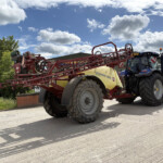 Blue tractor with red farming equipment attached, parked beside a building on a sunny day with scattered clouds