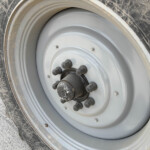 Close-up of a large vehicle wheel with a gray rim, lug nuts, and a muddy tire edge on a dusty surface
