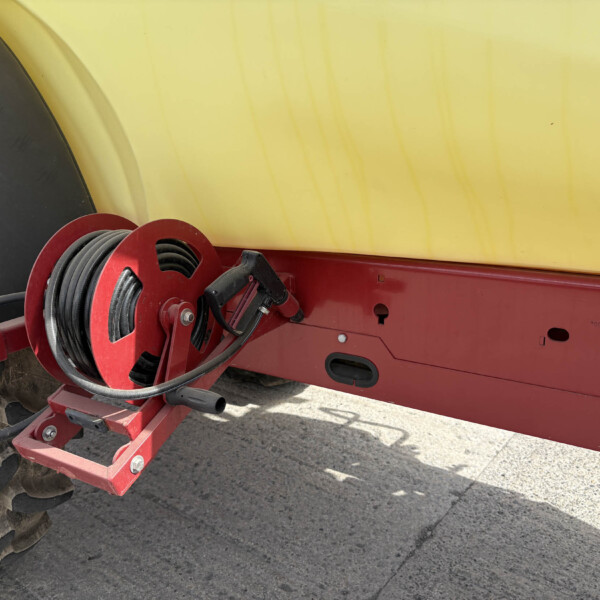 Red trailer frame with a black hose reel and coiled hose mounted beside a yellow panel/tank surface.