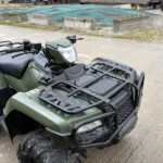 Green ATV (quad bike) with front rack and headlight parked on a dirt lot near an industrial yard.
