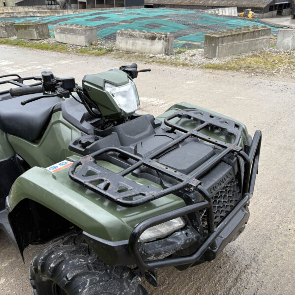 Green ATV (quad bike) with front rack and headlight parked on a dirt lot near an industrial yard.