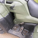 Olive-green Honda ATV side panel with logo, black seat, and footstep area visible outdoors.