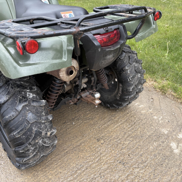 Rear view of a muddy green Honda ATV with a metal cargo rack, red taillight, and rugged off-road tires on a dirt path.