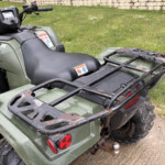 Rear view of a green Honda ATV with a rusted cargo rack and red taillights on a dirt surface.