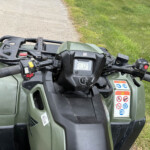 Front view of a green ATV's handlebar and dashboard, showing grips, controls, and a digital display with grassy area beside a dirt path in the background