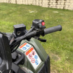 Close-up of a motorcycle/ATV handlebar with a red 4WD switch and warning sticker, grassy yard behind.