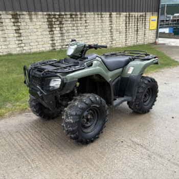 Green quad bike parked on a concrete path beside a brown stone wall and grassy patch under an industrial building backdrop