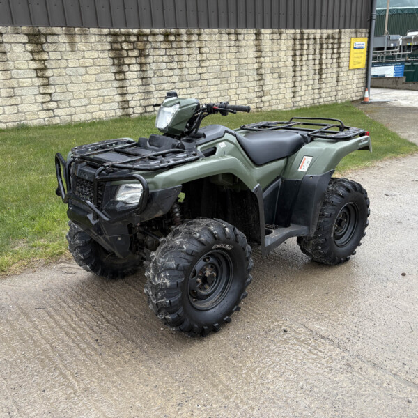 Green quad bike parked on a concrete path beside a brown stone wall and grassy patch under an industrial building backdrop