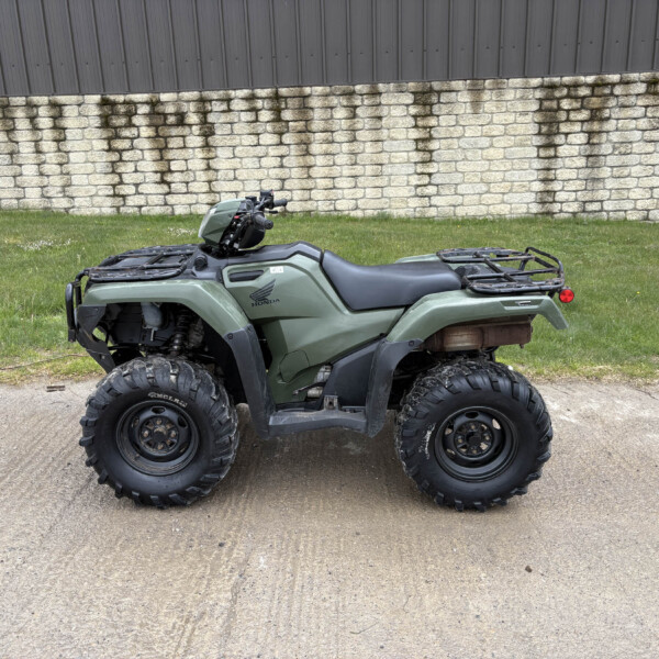Green Honda ATV parked on a dirt path with grass nearby and a stone wall behind it.