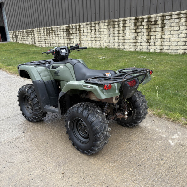 Green Honda ATV parked on a dirt/gravel path beside a grassy area and a stone retaining wall by a metal industrial building.