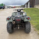 Rear view of a green ATV parked on a gravel lot with farm machinery and a large metal building in the background.