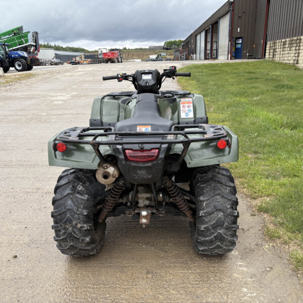 Rear view of a green ATV parked on a gravel lot with farm machinery and a large metal building in the background.